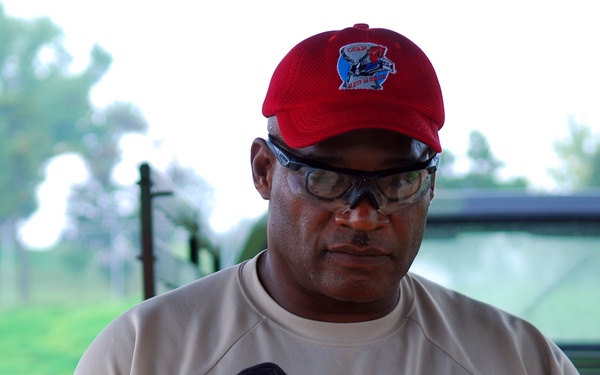 A man wearing a red baseball cap and safety glasses is working with lumber in what appears to be an outdoor or semi-covered workspace. He is wearing black work gloves and a tan/khaki athletic shirt while handling large wooden boards. A vehicle and green trees are visible in the blurred background.