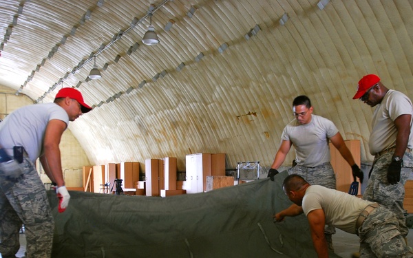 Five military personnel in U.S. Army/Air Force uniforms are working together inside a large inflatable or quonset-style structure to fold or unfold a large olive-drab canvas tarp or tent material. Three individuals are wearing red baseball caps, while others wear standard military camouflage uniforms. The background shows stacked cardboard boxes and wooden crates, suggesting a logistics or supply operation.