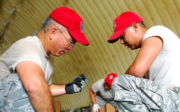Two military personnel wearing US Air Force Airman Battle Uniforms (ABU) and matching red caps are crouched down working with a large olive-drab military duffel bag inside what appears to be an inflatable or fabric-walled military tent structure. Both individuals are wearing black tactical gloves and combat boots. A third person in similar attire is partially visible in the background.