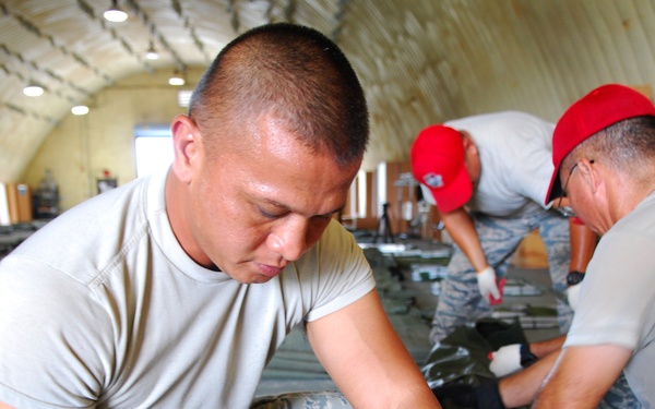 Military personnel in US Air Force ABU (Airman Battle Uniform) camouflage pants are working inside what appears to be a Quonset hut or similar military building. The foreground subject is kneeling and handling a large olive drab military duffel bag, while two other individuals in the background are similarly engaged with gear. The setting suggests a gear inspection, packing, or equipment accountability activity.