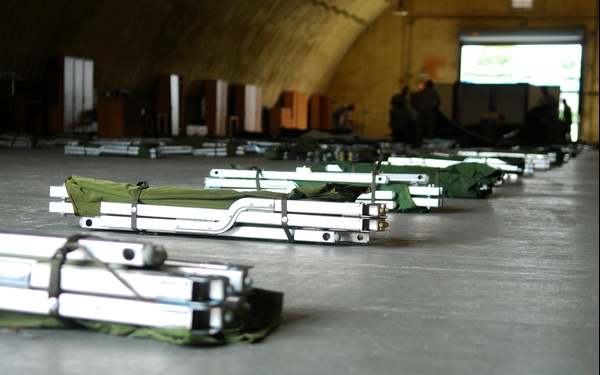 The image shows a large arched military or industrial building with numerous folded field cots or stretchers laid out in rows on the floor. The cots appear to be aluminum-framed with olive drab/green canvas fabric, suggesting military surplus or emergency preparedness equipment. Personnel are visible in the background near the open end of the structure.