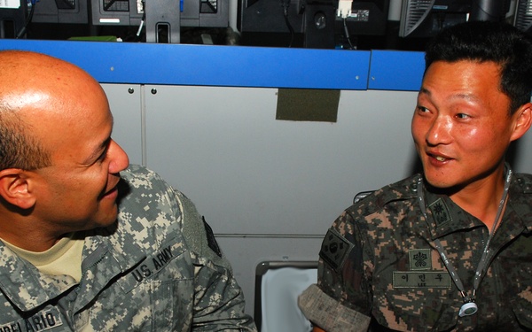 Two military personnel are engaged in conversation at what appears to be a command or operations center. One soldier wears a U.S. Army ACU (Army Combat Uniform) with the name tape 'CANDELARIO' and a Staff Sergeant chevron patch, while the other wears a Republic of Korea (ROK) Army camouflage uniform with Korean text on the name tape. A placard on the table reads '3rd BCD-K', suggesting a joint U.S.-Korean military exercise or liaison setting.