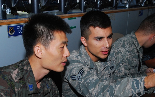 Two military personnel in different camouflage uniforms are seated together at a workstation, appearing to collaborate while looking at a computer monitor. The individual on the right wears a U.S. Air Force Airman Battle Uniform (ABU) with visible rank insignia (Senior Airman) and a name tape reading 'JACOBS'. The individual on the left wears what appears to be a Republic of Korea (ROK) military digital camouflage uniform, suggesting a joint or combined military exercise or operation.