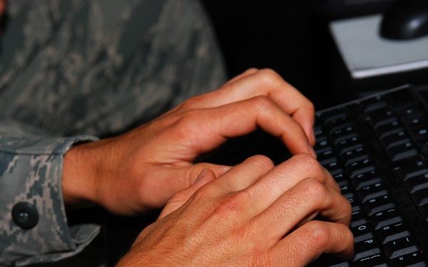 A person wearing military camouflage uniform (appears to be US Air Force ABU - Airman Battle Uniform pattern) is typing on a black computer keyboard. The image is a close-up shot focusing on the hands and keyboard, with a computer mouse visible in the upper right background. The setting appears to be an office or workstation environment.