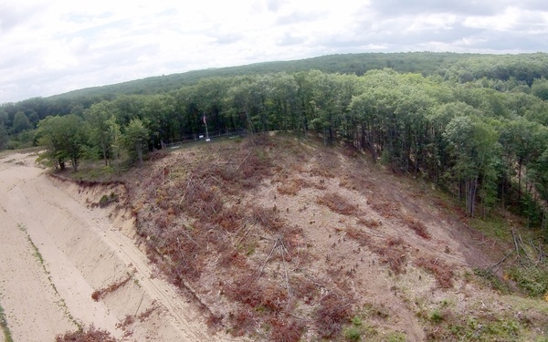An aerial drone photograph showing a large cleared and graded sandy area surrounded by dense deciduous and coniferous forest. The cleared zone contains scattered fallen trees, brush piles, and disturbed soil, indicating recent land clearing or logging activity. A road or path runs along the left edge of the cleared area, with what appears to be a small structure or facility visible in the far left.