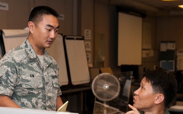 Two military personnel are engaged in a conversation in what appears to be a military operations or briefing room. One individual is standing and wearing US Air Force ABU (Airman Battle Uniform) with a name tape reading 'KIM', while the other is seated and wearing an olive green flight suit with squadron patches. They appear to be discussing work-related matters across a computer workstation.