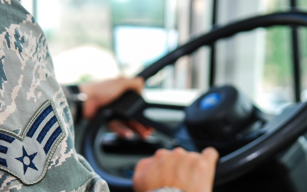 A U.S. Air Force enlisted member in Airman Battle Uniform (ABU) digital camouflage is seated at the wheel of a large vehicle, likely a military bus or transport vehicle. The rank insignia on the sleeve is clearly visible, identifying the individual as a Senior Airman (E-4). The image is a close-up shot emphasizing the uniform detail and driving activity.