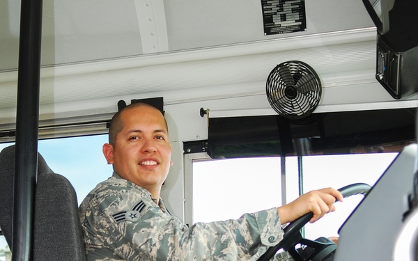 A U.S. Air Force enlisted airman in ABU (Airman Battle Uniform) camouflage is seated in the driver's seat of a large military or government vehicle, smiling at the camera. The individual appears to be an Airman First Class based on the rank insignia visible on the uniform. The vehicle appears to be a bus or large transport vehicle, as indicated by the 'TIRE CARRIER ACCESS' text visible at the bottom of the frame.