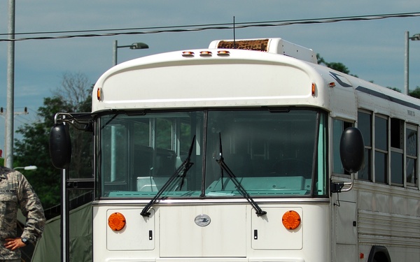 A U.S. Air Force service member in ABU (Airman Battle Uniform) camouflage stands beside a white Blue Bird military bus on a paved surface at what appears to be a military installation or field exercise area. The bus bears an Air Force license plate reading 'AF09B02752'. Military tents and equipment are visible in the background.