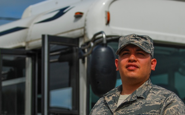A U.S. Air Force enlisted service member in Airman Battle Uniform (ABU) stands in front of a white passenger bus. The individual appears to be posing for an official or portrait-style photograph outdoors in daylight. The name tape and branch tape are partially visible on the uniform.