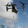 A military tiltrotor aircraft, identifiable as a Bell Boeing V-22 Osprey, is hovering over a forested area while a soldier rappels down a rope from the open fuselage door. The aircraft's rotors are in motion, visible through motion blur, and a red flag or marker is attached to the top of the fuselage. The scene depicts a military fast-rope or rappelling training or operational exercise.