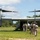 A military tiltrotor aircraft, identifiable as a Bell Boeing V-22 Osprey, is landed on a grassy clearing with its rotors spinning. Several military personnel in camouflage uniforms are approaching or boarding the aircraft through its rear ramp. The setting appears to be a wooded or jungle-adjacent landing zone under partly cloudy skies.