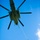 A large military helicopter is photographed from below against a clear blue sky, with multiple personnel suspended from a single rope hanging beneath the aircraft. The individuals appear to be in uniform and are clustered along the rope in what appears to be a Special Patrol Insertion/Extraction (SPIE) or similar helicopter extraction technique. The photo is taken from a low angle looking upward, providing a dramatic perspective of the operation.
