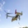 A military MV-22 Osprey tiltrotor aircraft hovers low over a grassy field while personnel conduct a fast-rope insertion exercise. At least one person is visible descending a rope from the open rear ramp of the aircraft, while ground personnel observe from below. The aircraft displays markings consistent with a U.S. Marine Corps unit, with a red tail fin marking and the number '8220' visible.
