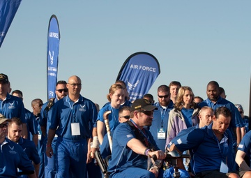 A group of Air Force Wounded Warriors (AFW2) participants are gathered at what appears to be an athletic or ceremonial event. The group includes individuals in wheelchairs, on mobility scooters, and walking, all wearing matching blue Air Force polo shirts. Multiple 'Wounded Warrior - Care Beyond Duty' and 'AFW2' branded banners are prominently displayed in the background.