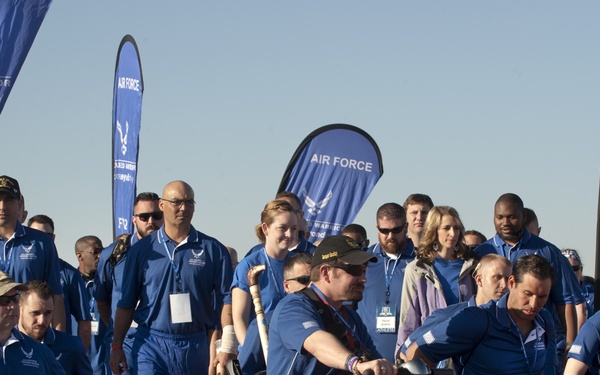 A group of Air Force Wounded Warriors (AFW2) participants are gathered at what appears to be an athletic or ceremonial event. The group includes individuals in wheelchairs, on mobility scooters, and walking, all wearing matching blue Air Force polo shirts. Multiple 'Wounded Warrior - Care Beyond Duty' and 'AFW2' branded banners are prominently displayed in the background.
