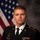 A formal U.S. Army official portrait of a soldier identified by nameplate as 'ROBERTS', wearing the Army Service Uniform (ASU) in black with gold trim. The subject is posed in front of a U.S. flag and a gray studio backdrop. The uniform displays several awards, badges, and insignia consistent with an enlisted or junior officer.