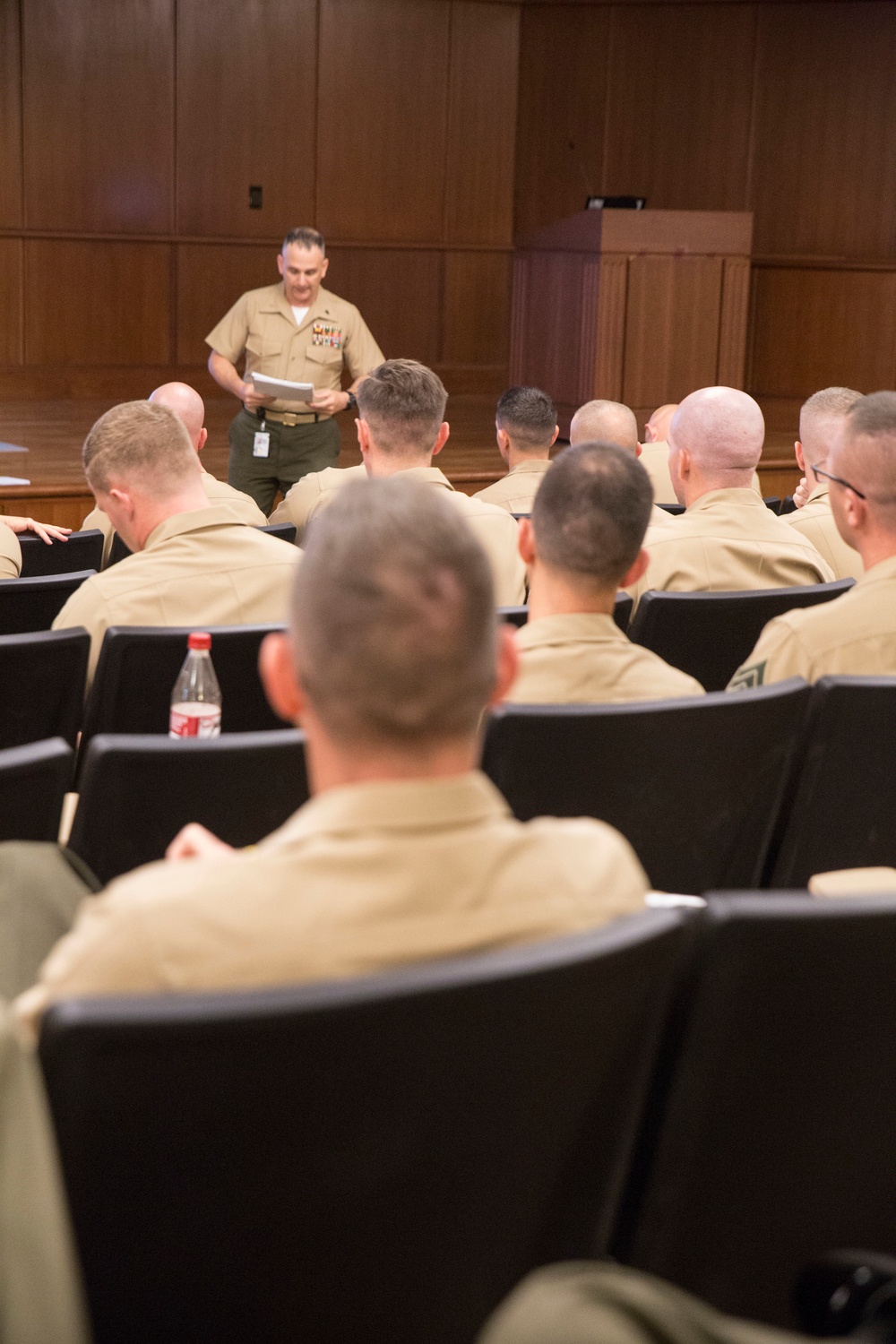 A military briefing or training session is taking place in a formal auditorium or conference room with wood-paneled walls. A senior military officer in tan/khaki uniform with ribbons and medals is standing at the front, reading from papers, while multiple service members seated in rows of black chairs face him. The setting and uniforms are consistent with United States Marine Corps (USMC) service charlies or similar garrison uniform.