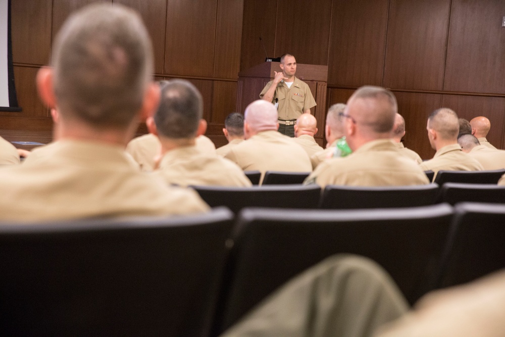 A military briefing or lecture is taking place in a formal auditorium or conference room. A single uniformed presenter stands at a podium addressing a large audience of seated military personnel, all wearing matching tan/khaki uniforms. The presenter appears to be making a hand gesture while speaking to the group.