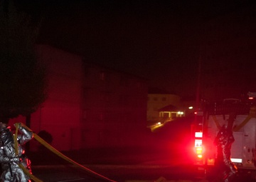 A nighttime emergency response scene showing personnel in protective suits handling a fire hose on a road. A fire truck with active red emergency lights is visible in the background. The scene appears to be a firefighting or hazmat operation conducted in darkness.