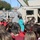 Two U.S. Army soldiers in ACU (Army Combat Uniform) are presenting a military HMMWV (Humvee) to a large group of elementary school children outdoors. One soldier is actively speaking and pointing while children raise their hands with questions. A female adult, likely a teacher, stands to the right supervising the students.