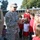 A U.S. Army soldier in ACU (Army Combat Uniform) is speaking to a group of elementary school children next to a military vehicle, likely a HMMWV (Humvee). The setting appears to be a school campus, suggesting this is an educational outreach or career day event. An adult woman in a red top, likely a teacher, supervises the children in the background.