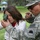 A U.S. Army soldier wearing CBRN (Chemical, Biological, Radiological, and Nuclear) unit insignia is instructing a young woman on how to use a military lensatic compass at what appears to be an outdoor public event. The soldier is pointing in a direction while the young woman holds the compass up to her eye to take a bearing. The setting appears to be a community or military family day event in a park.
