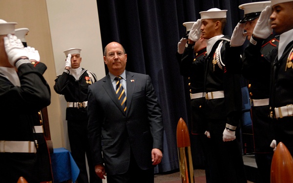 A civilian in a dark suit and striped tie walks along a red carpet through a formal military honor cordon. Uniformed service members from multiple branches, including the Navy and Marine Corps, stand at attention and render salutes on both sides of the aisle. The setting appears to be an official government or military ceremony room with decorative artillery shell stanchions lining the path.