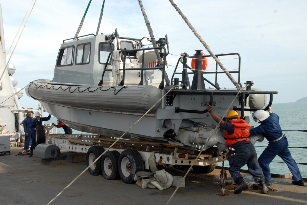 A gray rigid-hull inflatable boat (RHIB) is being lifted or positioned by crane onto a multi-axle trailer on what appears to be the deck of a military or coast guard vessel. Several personnel in uniform, safety vests, and hard hats are guiding and steadying the boat using lines and physical effort. The operation is taking place at sea, with open water and a distant landmass visible in the background.