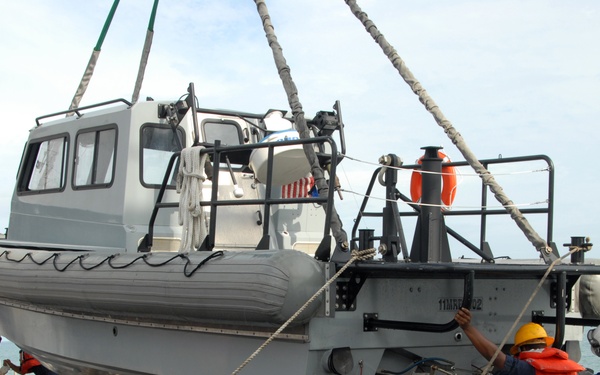 A gray rigid-hull inflatable boat (RHIB) is being lifted or positioned by crane onto a multi-axle trailer on what appears to be the deck of a military or coast guard vessel. Several personnel in uniform, safety vests, and hard hats are guiding and steadying the boat using lines and physical effort. The operation is taking place at sea, with open water and a distant landmass visible in the background.