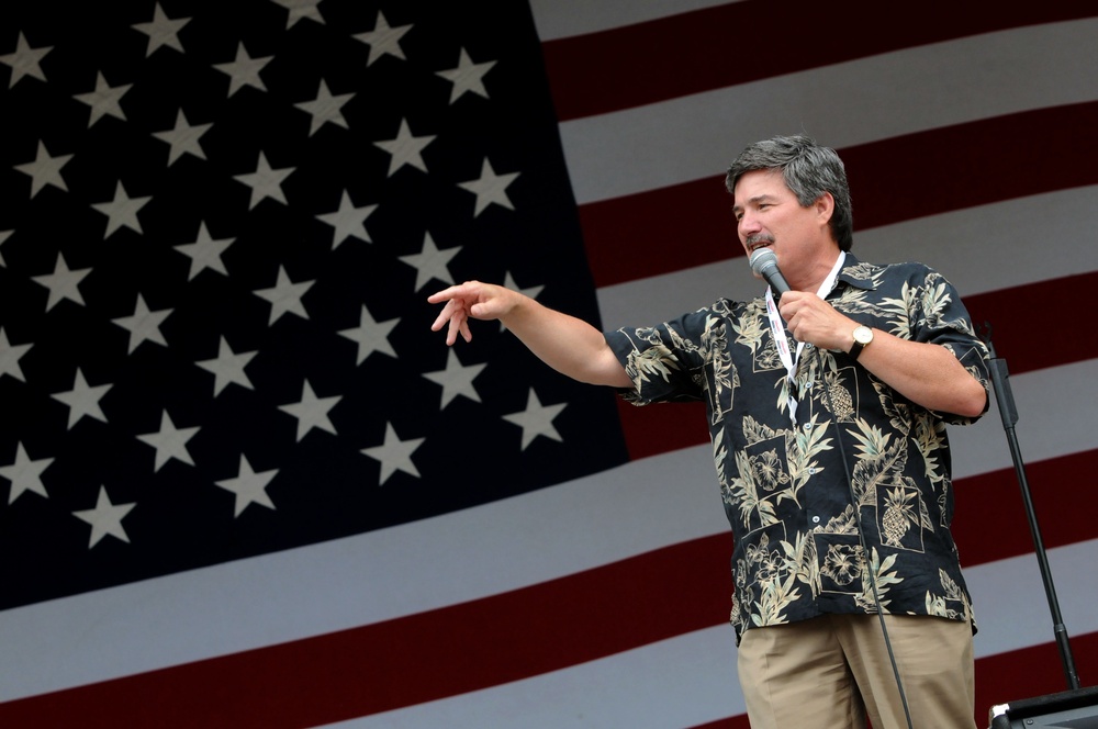 A middle-aged man with dark hair and a mustache is speaking into a microphone on a stage, with a large American flag displayed prominently in the background. He is wearing a dark floral Hawaiian-style shirt and khaki pants, gesturing with his left hand while holding the microphone with his right. The setting suggests a patriotic public event or rally.