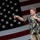 A middle-aged man with dark hair and a mustache is speaking into a microphone on a stage, with a large American flag displayed prominently in the background. He is wearing a dark floral Hawaiian-style shirt and khaki pants, gesturing with his left hand while holding the microphone with his right. The setting suggests a patriotic public event or rally.