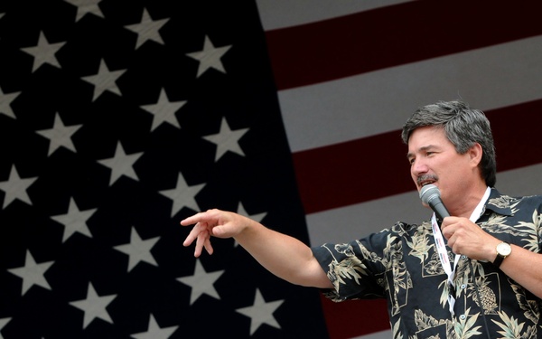 A middle-aged man with dark hair and a mustache is speaking into a microphone on a stage, with a large American flag displayed prominently in the background. He is wearing a dark floral Hawaiian-style shirt and khaki pants, gesturing with his left hand while holding the microphone with his right. The setting suggests a patriotic public event or rally.
