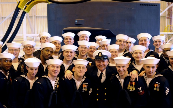 A large group of U.S. Navy personnel posing for a formal group photograph inside what appears to be a naval facility or hangar. The majority are enlisted sailors wearing standard Navy dress blue uniforms with white 'Dixie cup' covers, while one central figure appears to be a senior enlisted or officer wearing a peaked cap and dress uniform with medals. A dark-hulled submarine or vessel is visible in the background.