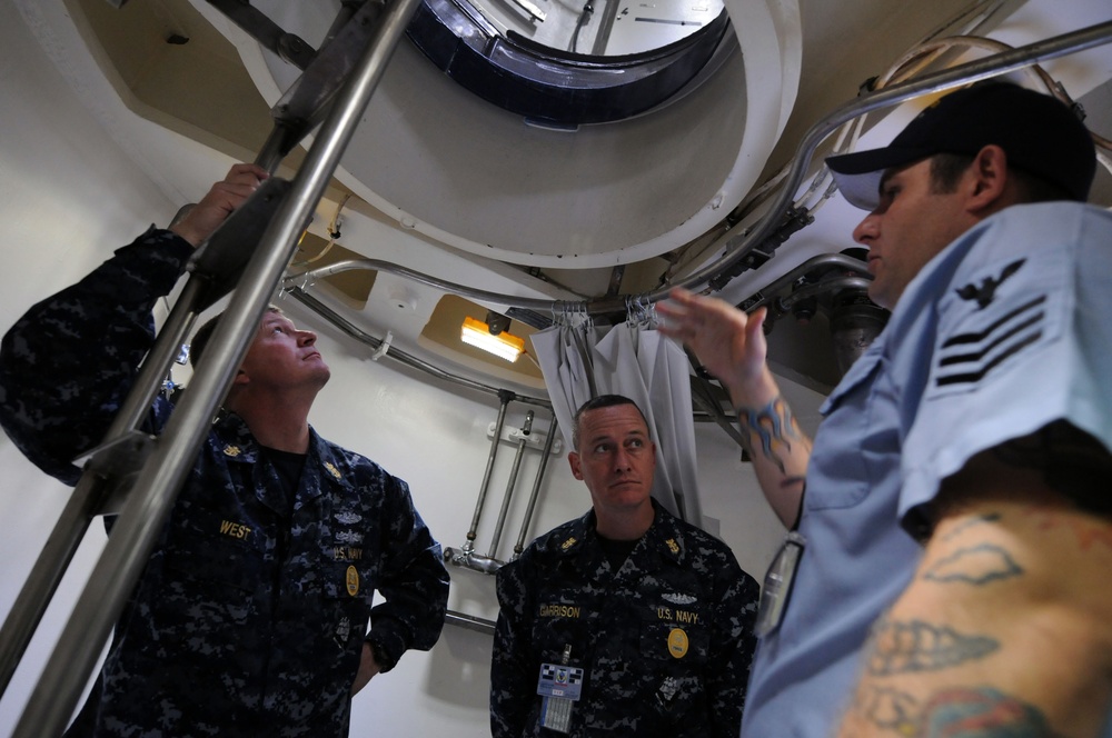 Three military personnel, two in U.S. Navy digital camouflage uniforms and one in a light blue uniform with a black cap, are examining an overhead circular hatch or opening inside what appears to be a naval vessel or facility. Name tapes visible on the camouflage uniforms read 'WEST' and 'GARRISON.' The person in the light blue uniform, who has visible tattoos on their arm, appears to be explaining or demonstrating something about the structure above.
