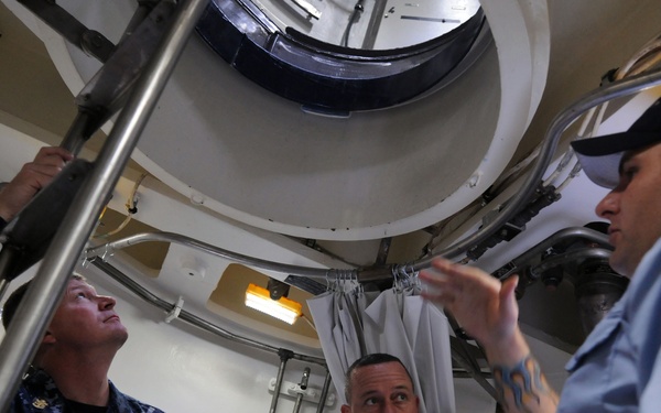 Three military personnel, two in U.S. Navy digital camouflage uniforms and one in a light blue uniform with a black cap, are examining an overhead circular hatch or opening inside what appears to be a naval vessel or facility. Name tapes visible on the camouflage uniforms read 'WEST' and 'GARRISON.' The person in the light blue uniform, who has visible tattoos on their arm, appears to be explaining or demonstrating something about the structure above.