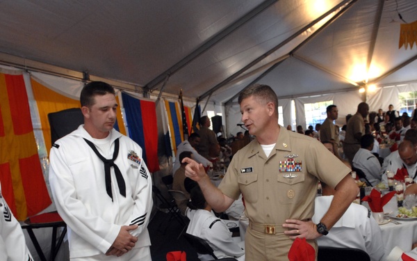 A formal military dining event taking place under a large tent, featuring U.S. Navy personnel in both white dress uniforms and khaki officer's uniform. A senior officer wearing ribbons and medals is engaged in conversation with enlisted sailors standing near a formally set dining table. The setting appears to be an official Navy luncheon or awards ceremony with decorative flags and bunting visible in the background.