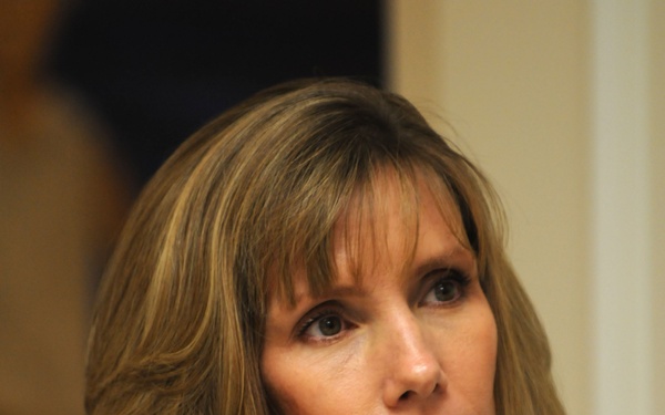 A middle-aged woman with shoulder-length brown hair with highlights is photographed in a close-up portrait style. She is wearing a gray cable-knit v-neck sweater and appears to be looking upward with a serious or concerned expression. The background is blurred, suggesting a shallow depth of field.