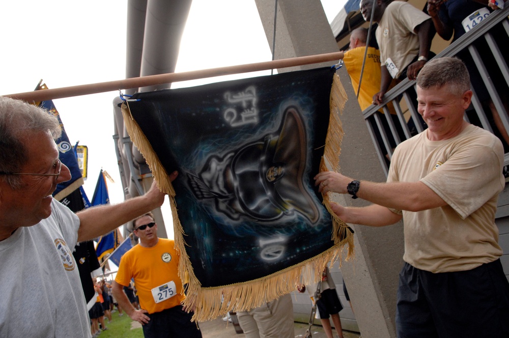 Two men are holding up a decorative banner or flag suspended from a wooden pole at what appears to be an outdoor event or race. The banner features dark airbrushed artwork depicting what appears to be a boot or shoe with wings and other design elements, with gold fringe along the bottom and sides. In the background, participants wearing race bibs and orange/yellow shirts are visible near stadium-like structures.