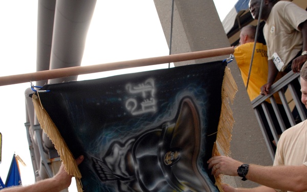 Two men are holding up a decorative banner or flag suspended from a wooden pole at what appears to be an outdoor event or race. The banner features dark airbrushed artwork depicting what appears to be a boot or shoe with wings and other design elements, with gold fringe along the bottom and sides. In the background, participants wearing race bibs and orange/yellow shirts are visible near stadium-like structures.