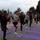 A large group of military personnel in physical training attire are performing yoga outdoors on purple and red yoga mats arranged in rows. Participants are executing a tree pose (Vrksasana) with hands in prayer position and one leg raised. The setting appears to be a military installation courtyard or parade ground, with trees and fencing visible in the background.
