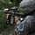 A U.S. Army soldier in ACU (Army Combat Uniform) digital camouflage is shown in a firing position, aiming an M4 carbine rifle with attached accessories. The soldier is photographed from behind and to the side in an outdoor, vegetated environment. A unit patch is visible on the left shoulder of the uniform.
