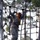 A soldier in US Army ACU (Army Combat Uniform) camouflage is navigating a rope obstacle course at what appears to be a military training facility. The soldier is wearing a safety harness with yellow and red straps, black gloves, and sunglasses while traversing a large rope net structure. A second soldier in similar uniform is visible in the background on the same obstacle.