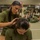 Two female military recruits in olive green t-shirts are seated on a barracks floor. One recruit is braiding or styling the wet hair of the other, who is seated in front reading a printed document. The background shows a military barracks bay with bunk beds, gear bags, and additional recruits in similar uniforms.