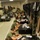 Female military recruits, dressed in olive green PT uniforms, are seated on the floor of a barracks bay writing in notebooks during what appears to be a free period or study time. The room contains a rifle rack with multiple rifles secured along the right wall, black footlockers used as desks, and bunk beds along the left wall. The scene is consistent with a U.S. Marine Corps recruit training depot environment.