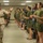 A female U.S. Marine drill instructor in desert MARPAT camouflage uniform and campaign cover is addressing a line of female recruits in an indoor barracks or training facility. The recruits are wearing olive green PT shorts and t-shirts, standing at attention while holding small bags or pouches. This appears to be a Marine Corps recruit training or boot camp scenario.