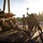 1st Cavalry arrives in Poland Military personnel in camouflage uniforms are working to secure or unsecure a piece of heavy military equipment on a wooden platform or ramp. One soldier is wielding a large sledgehammer, suggesting they are driving or removing securing pins or stakes. A civilian photographer in a red jacket is documenting the activity from the right side.