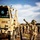 1st Cavalry arrives in Poland U.S. Army soldiers are conducting a military vehicle rail-loading operation, with a tactical wheeled vehicle marked 'CCP 12' being guided onto a flatcar by a ground guide giving a thumbs-up signal. The driver's placard reads 'SGT KING,' and the directing soldier wears a yellow 'CAV' identifier tab. Multiple soldiers in multicam uniforms and some wearing high-visibility vests are visible in the background managing the convoy.