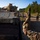 1st Cavalry arrives in Poland Military personnel in camouflage uniforms are directing a tracked armored fighting vehicle, identified as an M2 Bradley Infantry Fighting Vehicle, onto or off of a flatbed rail car. A soldier in the foreground is using hand signals to guide the vehicle operator. The scene appears to be a rail loading or offloading operation in a wooded area.
