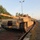 1st Cavalry arrives in Poland Multiple M1 Abrams main battle tanks in desert tan livery are loaded onto flatbed railway cars, suggesting a military logistics or transport operation. The lead tank bears the marking 'E31' on its turret. The setting appears to be a rail yard or loading facility flanked by trees and street lighting, photographed during early morning or late afternoon light.